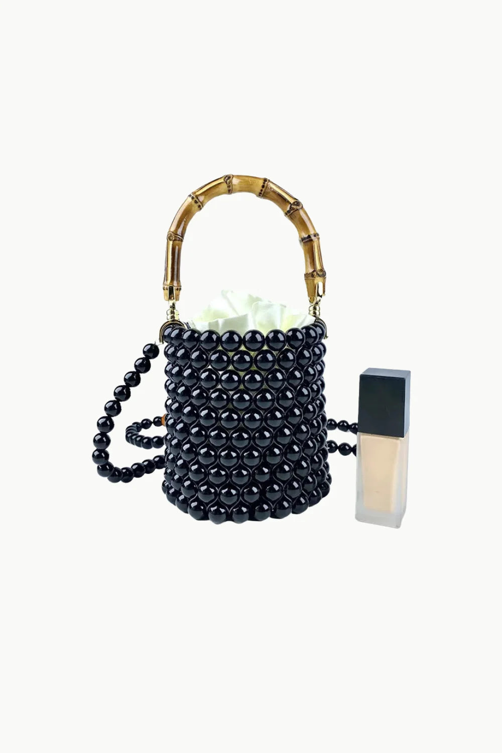 Black beaded bucket bag with bamboo handle and shoulder strap next to liquid foundation
